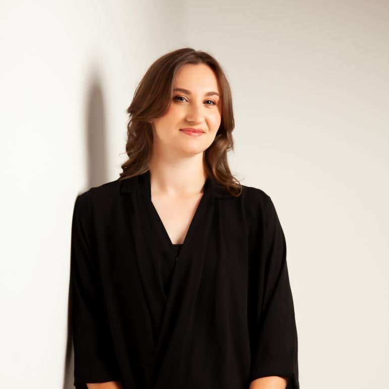 An image of Amy Arthur. Amy is a white woman with brown hair that has been curled. She wears a black blouse and leans against a white wall.